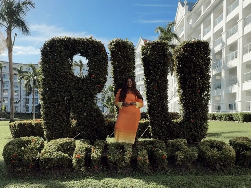 Solo traveler standing in front of the RIU Ocho Rios hedge sign with the hotel resort in the background.